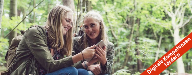 Zwei junge Frauen sitzen in einem Wald mit einem Smartphone.