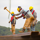 Drei Menschen mit Arbeitskleidung und Helmen stehen auf einem Stahlträger und arbeiten. 