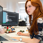 Ein Frau mit roten Haaren sitzt vor zwei Computern, auf denen etwas in Codiersprache angegeben ist. 