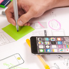 Für das Lernen im Unterricht können Smartphones oder Tablets ein nützliches Werkzeug sein. Smartphone auf dem Schreibtisch, daneben die Hand einer Person, die etwas mit einem Stift notiert