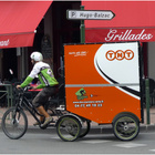 Das Bild zeigt einen Fahrradkurier auf einem Lastenrad. Hinten dran hat er einen orangenen Kasten, der auf Rädern fährt.