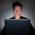 Mann sitzt in dunklem Raum vor dem Laptop, sein Gesicht wird durch das Bildschirmlicht beleuchtet. 