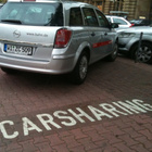 Ein graues Auto parkt auf einem Parkplatz und vor ihm auf dem Boden steht in Großbuchstaben "Carsharing". 