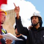 Ein Mann mit einem weißen Helm deutet auf einen Punkt in der Ferne, ein zweiter Mann steht daneben und hat einen Bauplan in der Hand. 