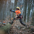 Ein Mann in Schutzausrüstung steht auf zwei Baumstämmen im Wald und bearbeitet den einen mit einer Maschine. 