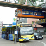 Schwebebahn fährt oberhalb einer Straße entlang; darunter ein haltender Bus und ein haltendes Auto