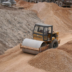 Eine Walzmaschine fährt auf einem Kiesweg, drumherum sind Berge aus Sand und Kies zu sehen. 