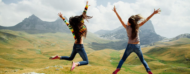 Zwei Mädchen springen in die Luft, im Hintergrund eine Berglandschaft