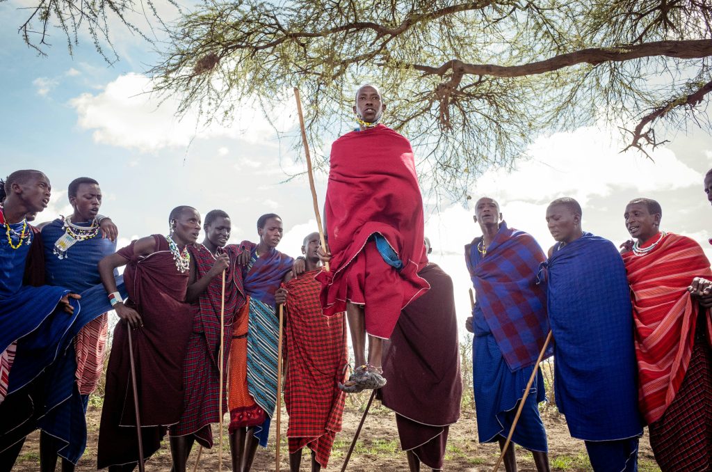 Gruppe von Maasai-Männern in traditioneller Kleidung, einer springt hoch, umgeben von Bäumen und offenem Himmel