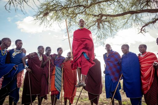 Gruppe von Maasai-Männern in traditioneller Kleidung, einer springt hoch, umgeben von Bäumen und offenem Himmel