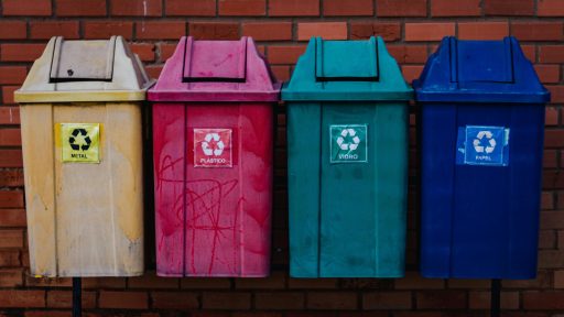 Vier Mülltonnen für Recycling vor roter Backsteinwand, beschriftet mit 'Metal', 'Plastik, 'Glass' und 'Papier'