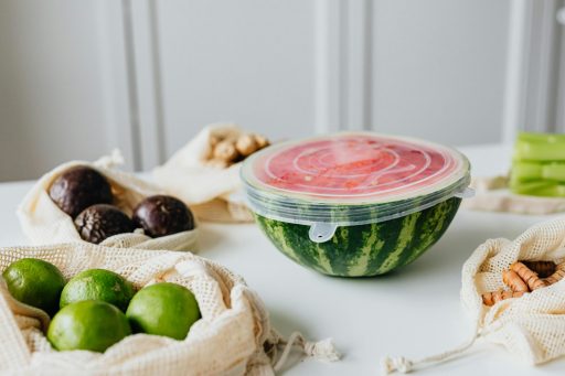 Halbierte Wassermelone in einer durchsichtigen Plastikschüssel mit Deckel auf einem weißen Tisch, umgeben von Avocados, Limetten und Nüssen in Stoffbeuteln