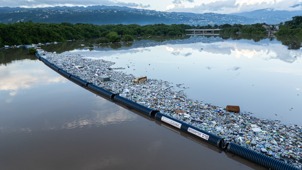 Schwimmende Barriere sammelt Plastikmüll auf ruhigem Gewässer vor bewaldetem Ufer und Bergen im Hintergrund