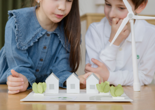 Zwei Kinder schauen auf einen Tisch mit Häusern aus Papier.