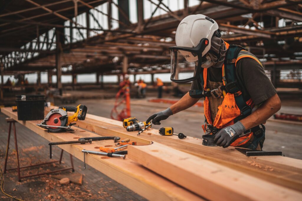 Ein Handwerker steht in einer Lagerhalle mit Helm und Handschuhen vor einem langen Holzbrett und vielen Werkzeugen.