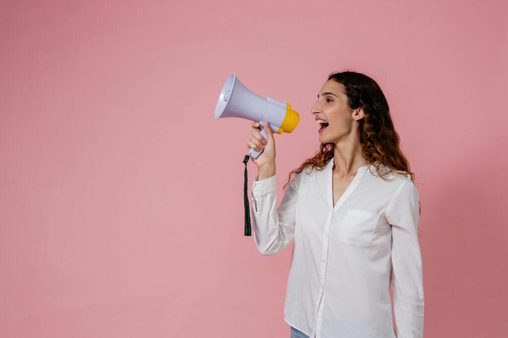 Eine Frau spricht in ein einem Megaphone und steht vor einer pinken Wand.