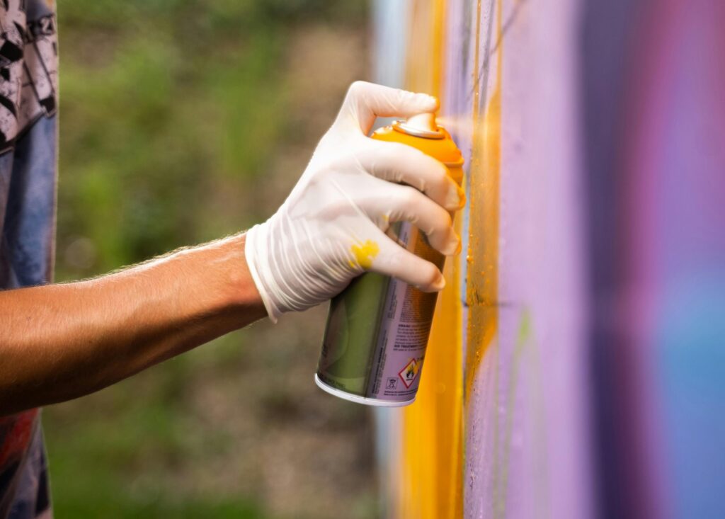Eine Hand mit Einmalhandschuhe sprüht mit einer Spraydose Farbe auf eine bunte Wand.