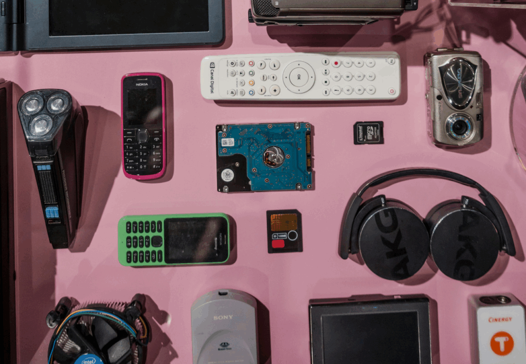 Verschiedene elektronische Geräte und Zubehörteile wie Mobiltelefone, Fernbedienung, Kopfhörer, Festplatte und Speicherkarten auf rosa Untergrund angeordnet