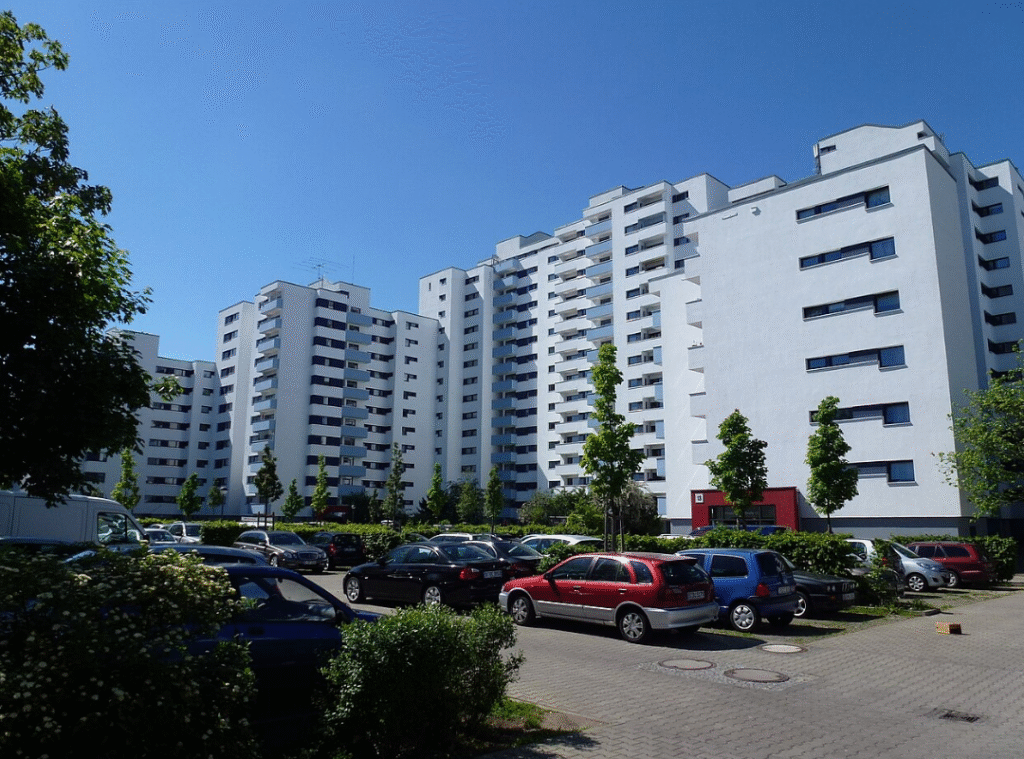 Ein Neubauwohnviertel mit weißen hohen Gebäuden. Davor ist ein Parkplatz.