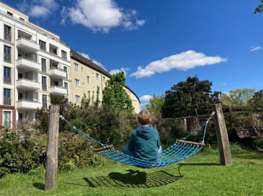 Ein Kind sitzt auf einer Hängematte im Schneidersitz auf einer Stadtgrünfläche. Im Hingergrund sind Neu-und Altbauten.